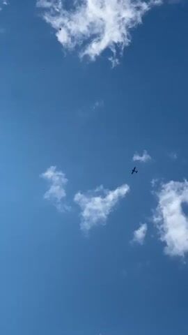 A small airplane flies across a bright blue sky dotted with fluffy white clouds. The airplane is dark in silhouette against the sky.