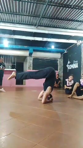 In an indoor dance or practice studio located in El Dátil, Venezuela, a person is performing an inverted balance, likely a headstand, on a brown-tiled floor. Dressed in black pants and a dark top, the individual balances on their head and forearms with legs extended vertically towards the corrugated metal ceiling.

Several other people are seated on the floor in the background, observing the activity. The room features multi-colored walls (white, blue, dark) and is illuminated by fluorescent tube lights. A counter or bar area and a window/door with metal bars are visible in the background. On a dark wall, a white logo and text reads "Danzes ROMINA" (possibly "Danzas Romina" or "Danza Romina"), accompanied by a dancer's silhouette. The scene suggests a focused practice or training session within a functional, no-frills environment.