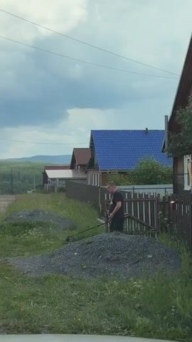 A man wearing glasses, a black t-shirt with an Adidas logo, and dark pants is operating a weed trimmer in front of a fence. The weed trimmer has a black handle and a yellow attachment. The man is standing next to a pile of gravel. In the background, there are several houses with different colored roofs and a cloudy sky. The environment appears to be a rural or suburban area with overgrown grass.