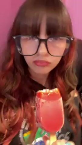 A woman is eating a red popsicle on camera. The woman has brown hair with red highlights and is wearing large, black-rimmed glasses. Her fingers are adorned with multiple rings, one with a blue stone. Her shirt is pink. She is licking and biting the popsicle. The background is a pink wall.