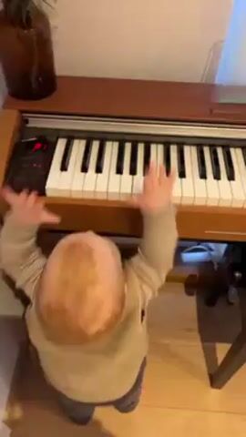 A baby is standing in front of a small electric piano, facing the instrument. The baby is wearing a light brown sweater, grey pants and has reddish hair. The baby is reaching out with both hands toward the piano keys, preparing to press them. The piano is a light brown wood-grain color. There is a brown glass bottle with green plants on top of the piano. The room is fairly brightly lit. A window with a white curtain is visible in the upper right. A chair with dark brown legs is partially visible under the piano.