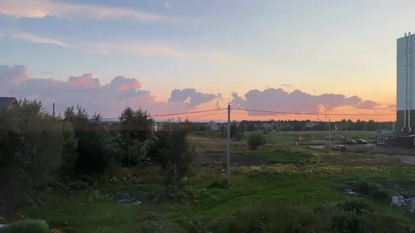 This media file captures a serene sunset scene in Vylgort, Russia. The sky is a canvas of soft hues, transitioning from a pale blue at the top to warm oranges and pinks near the horizon. Several fluffy clouds are illuminated by the setting sun, adding a touch of dramatic color to the sky.

The foreground is dominated by lush green grass and various trees and bushes, suggesting an undeveloped or rural area. A prominent utility pole with wires crosses the frame, a common sight in such landscapes. In the middle ground, a small settlement of houses can be seen, hinting at a nearby community. Further to the right, a modern, multi-story apartment building stands out, its beige and brown facade contrasting with the natural setting. A few cars are parked near the building, and a playground structure is visible in the distance.

The time of day is clearly evening, as indicated by the sunset and the soft, warm light bathing the scene. The weather appears clear and pleasant. The overall emotion conveyed is one of tranquility and peacefulness, with nature's beauty on full display. There is no visible text in the media file.