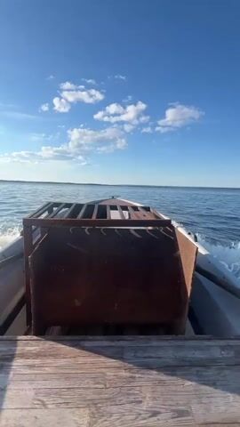 The video shows a person on a boat moving through calm water under a bright blue sky with scattered white clouds. The boat appears to be a small, possibly metal vessel with a wooden deck and a rusty metal structure in the foreground. The water is creating a wake behind the boat. In the distance, another boat is visible, and the scene is framed by a tree-lined shoreline. The sun is shining brightly, casting a glare on the water. The overall mood is peaceful and serene, suggesting a pleasant day on the water. The time of day appears to be late morning or early afternoon due to the brightness of the sun and the quality of the light. The location is identified as Siukterka, Russia. There is no visible text in the video.