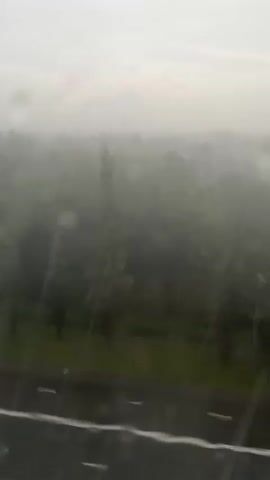 The video shows a hazy, overcast sky with a distant treeline and a road in the foreground. Raindrops are visible on the glass through which the video is taken, blurring the view and creating streaks. The overall impression is one of a gloomy, rainy day.