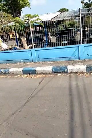 The video shows a view from a moving vehicle. The camera pans across a street scene. In the foreground, there is asphalt with some fallen leaves. A blue concrete barrier with a black and white striped curb runs along the side of the road. Behind the barrier is a metal fence, and behind the fence, there are trees and some buildings. A red and white flag is visible on a flagpole. There are also some people and motorcycles visible behind the fence. The overall impression is an outdoor scene on a sunny day.