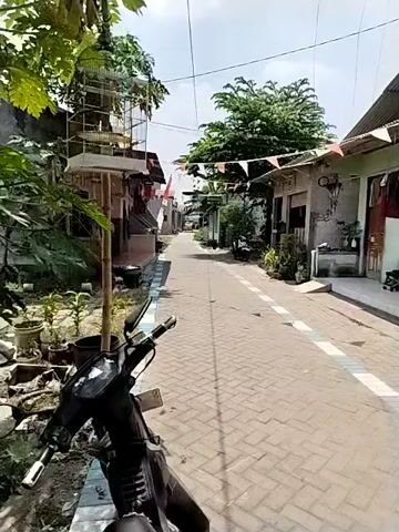 A motorcycle is parked on the side of a street paved with brick. The street is lined with houses and trees. Flags are strung across the street. The houses appear to be simple structures with corrugated metal roofs. The sky is blue and there are some clouds. The video is shot from a first-person perspective, presumably from the perspective of the person riding the motorcycle. The video appears to be taken during the day.