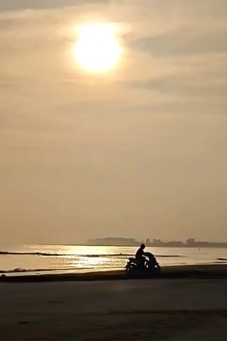 The video shows a beach scene with a bright sun shining through a hazy sky. In the foreground, there's a sandy beach. Two people are riding motorcycles along the water's edge. The person on the right rides toward the right side of the frame. The other one starts riding slowly in the opposite direction. In the distance, there's a landmass visible across the water. The overall tone of the video is serene, with the sun creating a warm, golden glow.