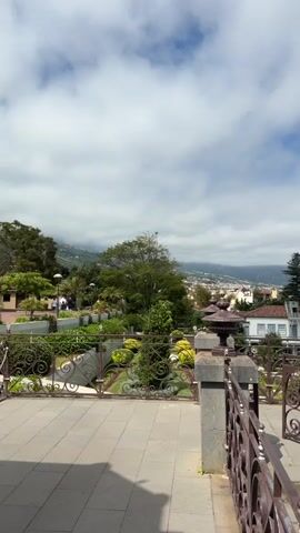 The video shows a panoramic view of a city and the ocean from a high vantage point, likely a balcony or terrace. In the foreground, there is a paved area with a decorative metal railing and stone pillars. Steps lead down into a well-maintained garden with manicured hedges, colorful flowers, and a circular fountain. Beyond the garden, the city stretches out with buildings featuring red-tiled roofs. In the distance, the blue ocean meets the horizon under a partly cloudy sky. A lone person is visible walking down the steps in the garden. The overall impression is of a scenic overlook in a coastal town on a bright, sunny day.
