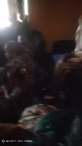 The video appears to be taken inside a dimly lit room. The camera is pointed at a cluttered area with various objects piled on top of each other, including patterned blankets and what looks like clothing. There is a person in the background, partially obscured, wearing a white head covering, possibly a hijab, seated on what seems to be a dark-colored sofa. The lighting is poor, making it difficult to discern details. There are sounds of children chattering.