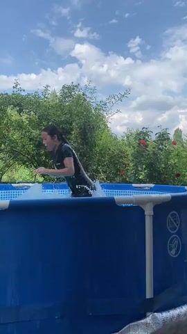 The video shows a young person in a black t-shirt and shorts jumping into a blue inflatable swimming pool. The pool is set up outdoors, with lush green trees and bushes, some of which have red flowers, in the background. The sky is blue with white, fluffy clouds, suggesting it is daytime and the weather is pleasant. The person emerges from the water, splashing and smiling, indicating they are enjoying themselves. The overall scene suggests a fun summer day in a backyard or garden setting.

There are some warning symbols visible on the side of the pool, including what appears to be a "no diving" symbol. The setting appears to be rural, possibly a village in Kyrgyzstan, given the context.