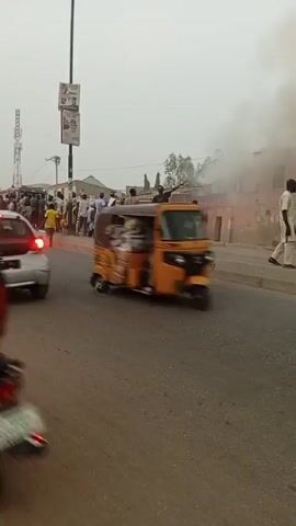 The scene shows a street with a crowd of people gathered on one side of the road near a building with smoke rising from it, presumably a fire. A fire truck with flashing lights is visible among the crowd. People appear to be observing the situation, and one individual is carrying something on their head away from the building. Motorbikes and a car pass on the road in the foreground. The sky is overcast, and there are poles with posters on them along the street.