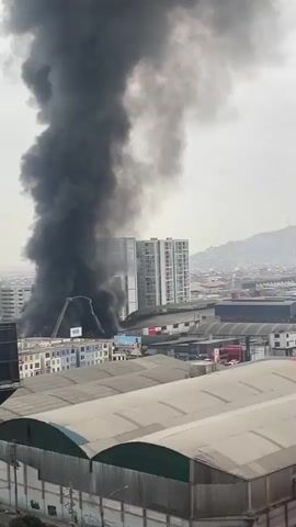A tall plume of thick black smoke billows into the sky from a burning building. The building appears to be a multi-story structure with a modern glass facade, surrounded by other buildings and industrial structures. In the foreground, there are large, flat-roofed warehouses. In the background, a hilly landscape is visible with more buildings. A fire truck ladder can be seen extended towards the burning building. There are no people clearly visible in the frame. The sky is overcast.