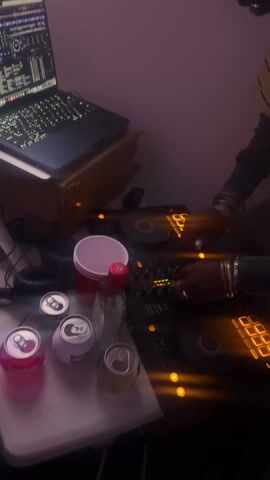 The video shows a blurry scene centered around a DJ setup.  On a surface, there are four cans of drinks (one pink), a red plastic cup, and a small bottle. Above this, a laptop sits on a wooden stand, its screen displaying DJ software. A person wearing a yellow plaid shirt and black sleeves is operating a DJ mixer. The lighting is dim with a purple hue, and there are illuminated buttons and controls on the mixer. The overall impression is that of a casual, possibly home-based, DJing session.  Sounds of a party can be heard.