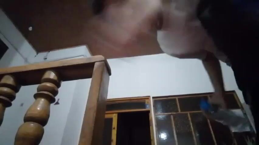 A boy in a white shirt and dark pants stands inside, near the top of a wooden staircase. He tosses a water bottle into the air so that it lands upright on the ledge of the staircase. The boy smiles and then begins to dance.