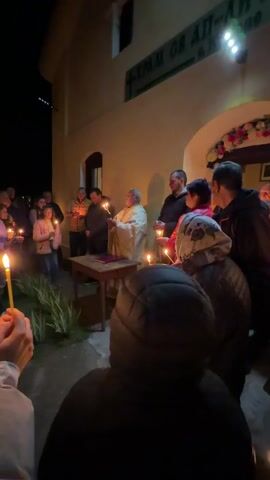 A religious ceremony is taking place at night outside a building. A priest stands behind a wooden table with a book on it, holding a cross adorned with lit candles. He is surrounded by a group of people holding lit candles. The building has a sign in a different language, decorated with flowers above the entrance. The scene is illuminated by the candle light, the building’s exterior lights, and some light from an unknown source off screen. The individuals are dressed in casual clothing and are standing near the entrance.