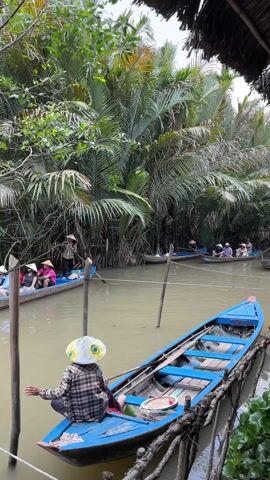The video shows a river scene in what appears to be Southeast Asia. The river is murky, with several blue boats carrying people. The people are wearing conical hats, a common sight in Southeast Asia. Lush green vegetation, including palm-like trees, lines the riverbanks. One blue boat is tied to wooden posts, with a person sitting in it, wearing a conical hat and a plaid shirt. The person appears to be holding onto the post. Other boats are moving along the river. There is a structure visible in the top right corner, possibly a thatched roof hut. The overall scene suggests a tourist attraction involving boat rides through the river. There are sounds of people speaking in the background, possibly market vendors.