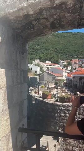 The video shows a view through an arched stone opening looking out onto a coastal town. In the foreground, on the right, a person's shoulder and back are visible, with long, wavy, light brown hair. They are holding a smartphone in their right hand, with their left hand near the phone. The person is looking down and slightly to the right. The view beyond the opening reveals a stone wall in the mid-ground, with an outdoor seating area that includes tables and potted plants. Beyond that, buildings with red-tiled roofs are clustered together. Further in the background, a lush green, tree-covered hillside rises steeply. To the left of the town, a rocky coastline meets the bright turquoise water of the sea. The sky is clear and blue. The overall impression is a sunny day in a picturesque Mediterranean or coastal location.