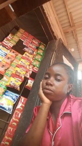 A woman in a pink shirt is standing in front of a wall of packaged snacks. She rests her face on her hand and looks at the camera. The snacks appear to be arranged on shelves. There is a lightbulb visible in the background. The camera angle is from a low position, looking up at the woman. The setting appears to be a small shop or kiosk. The woman appears calm and contemplative.