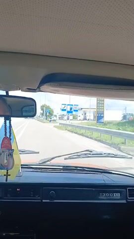 The video is filmed from the interior of a car, looking out the windshield. The dashboard is visible in the foreground, with the inscription "1300 S". A sun visor is overhead. The car is moving on a road. On the right side of the road, there is a gas station with pumps and signs. The gas station canopy is yellow. There are trucks parked at the gas station. Signs at the gas station are visible, including one that says "UKRNAFTA". There are also signs with prices. The road has white lines and a metal barrier on the right. There is greenery on the side of the road. The sky is cloudy. There is a blue and yellow flag attached to the car's windshield on the left side.