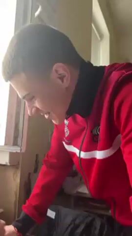 A young man in a red and white tracksuit is standing at a table near a window. He is looking down at a cellphone on the table and appears to be focused on what is displayed on the screen. Another young person is also looking at the phone, though their face is not fully visible. The phone is on top of a notebook and some other items. There are distracting noises in the background. A building is visible through the window.