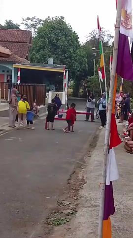 A street scene with a group of people, primarily children, participating in an outdoor event.  There is a red-colored obstacle, possibly for a race or game, set up on the asphalt road.  Several flags, including red and white ones, are visible, attached to poles on the right side of the frame and possibly in the background.  On the left, there are houses and a fence.  Adults are observing and supervising the children. The environment appears to be a residential area.