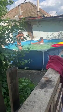 Two children are in an inflatable pool. One child is sitting on a white float. The other child is standing on the edge of the pool. There are inflatable toys in the pool. The pool is in a backyard with a house and trees.