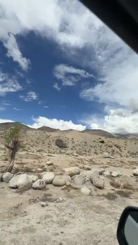 This is a point-of-view video captured from inside a vehicle traveling on a road through a mountainous, arid landscape. The sky is partly cloudy with scattered white clouds against a bright blue backdrop. The terrain is rocky and barren, with sparse vegetation. Large boulders are scattered across the landscape, and the mountains in the distance are rugged and brown. The road ahead is paved and appears to be in good condition. A yellow sign is visible on the side of the road, and a white car is seen in the distance. The interior of the vehicle is visible in the foreground, including the dashboard, windshield, side mirror, and window frame. A sticker with text and a phone number is affixed to the windshield. The overall impression is of a scenic drive through a remote, mountainous region.