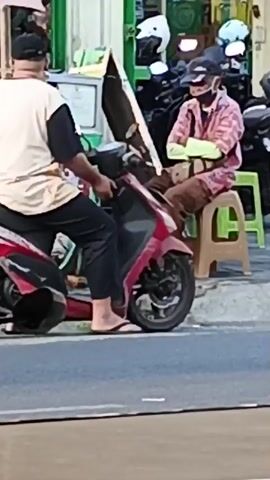 A man in a light-colored t-shirt and a dark cap is sitting on a red scooter. Another man in a patterned pink shirt, a black cap, and a face mask is standing next to the scooter, holding a bag. The scooter is parked on the side of a street. In the background, there is a stall with various items displayed, including helmets and what appears to be food. Another scooter is partially visible on the right. There is also a plastic chair visible. A car is passing by in the foreground, partially obscuring the view of the people and the scooter. The overall scene appears to be a street market or a roadside stall.