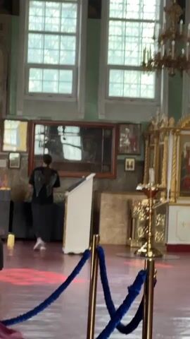 This media file captures the interior of an Orthodox church, likely in Nogornyi, Russia. The scene is dominated by religious iconography and ornate gilded objects, typical of Eastern Orthodox places of worship. A woman, dressed in a black t-shirt with a wing design and a patterned skirt, stands with her back to the camera, facing a large, framed mirror. She appears to be observing something within the mirror or the space it reflects.

The church interior features high arched windows with decorative grilles, allowing natural light to filter in. A large, multi-tiered chandelier hangs from the ceiling, hinting at a grander interior. The walls are painted a muted green, adorned with various religious paintings and icons. In the foreground, a velvet rope barrier with brass stanchions guides visitors, suggesting a designated path or area.

The focal point of the right side of the frame is an iconostasis, a wall of icons and religious imagery, beautifully decorated with gold accents. Lit candles are visible on ornate brass candle holders, indicating ongoing religious activity or a memorial. Other religious artifacts, such as chalices and crosses, are also present. The floor is a deep red, possibly linoleum or painted concrete.

The overall atmosphere seems serene and contemplative, with the soft lighting and the presence of religious artifacts creating a solemn mood. The woman's posture suggests introspection or prayer. The time of day appears to be daytime, given the natural light streaming through the windows. There is no visible text in the media file.