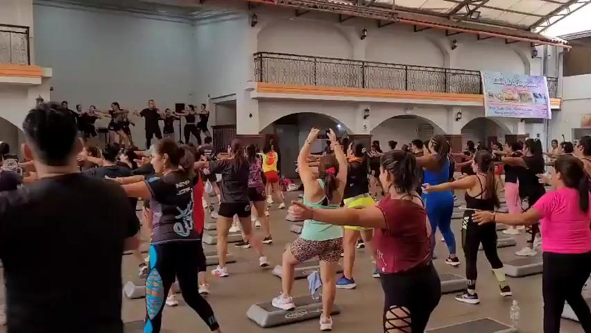 A group of people are participating in a step aerobics class in an indoor space.  The participants, predominantly women, are dressed in athletic wear and are positioned on aerobic steps.  A male instructor and several other individuals are leading the class from the front. The environment appears to be a large hall with arched windows and a balcony overlooking the activity. The participants are engaged in synchronized movements, following the instructor's lead.