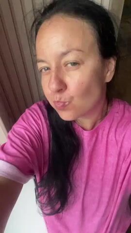 A woman is shown in a close-up shot, wearing a pink t-shirt and leopard-print pants. She is by a window with wooden blinds. In the first few seconds, she winks and puckers her lips at the camera. She then picks up a plastic container of food and a fork, and begins to eat. The video continues to show her eating for the remainder of the clip.