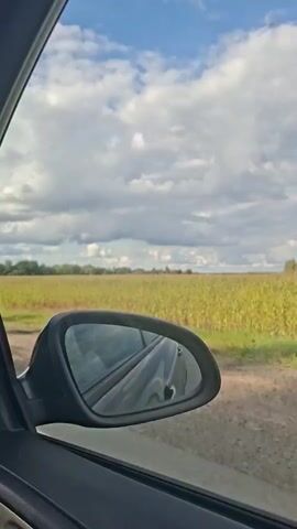 The video shows a view from inside a car, looking out the side window. A side-view mirror is prominent in the foreground, reflecting the interior of the car and the sky. Outside the window, there is a dirt road next to a large field of crops, likely corn, under a partly cloudy sky. The camera is panning slightly, giving a sense of movement as if the car is driving. The focus is on the interplay between the car's interior reflected in the mirror and the passing landscape.