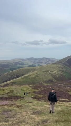 The video shows a scenic view of rolling green hills under a cloudy sky. In the foreground, a person wearing a backpack is walking away from the camera down a grassy path. Further down the path, two other people are walking in the distance. The landscape is characterized by undulating hills covered in green grass and patches of purple heather. In the far distance, a body of water and more distant hills can be seen, with a hazy horizon. The overall impression is of a natural, open landscape, likely a hiking trail.