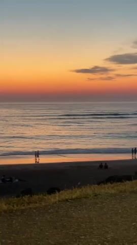 A tranquil beach scene unfolds at sunset. The sky displays a gradient of warm oranges and soft blues, meeting the calm ocean with gentle waves. Silhouettes of people are scattered across the dark sand. Near the water's edge, a small group stands together, some appearing to be conversing. Further down the beach, a single figure walks along the shoreline. In the foreground, dry grass and a dirt path are visible, framing the view of the serene coastal landscape.