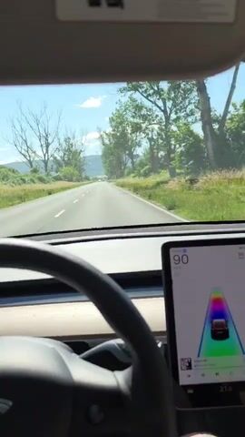 The video presents a first-person perspective from the driver's seat of a car, looking out through the windshield onto a road. In the foreground, the top part of a black steering wheel is visible on the left. To the right, a large, vertical digital display shows a speedometer indicating "90" and a graphic interface depicting a car icon on a road with a vibrant rainbow trail. Beyond the windshield, a two-lane asphalt road extends straight into the distance under a clear, bright blue sky with scattered white clouds. The road is bordered on both sides by green grassy areas and numerous trees, some with full green foliage and others with bare branches. Faint, distant hills or mountains are visible on the horizon. The car appears to be driving forward along the road.