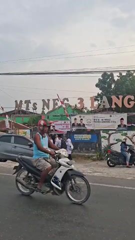 The video shows the entrance to a school with a gray car parked on the side of the road. Several students in uniforms are walking near the school entrance. One student is walking away from the camera, and others are approaching the school. A person is standing on the left side of the road, facing away from the camera. There are Indonesian flags displayed at the entrance. The sky appears overcast.