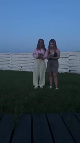 Two young women stand on a lawn in front of a white picket fence on a clear day. One is wearing a light pink jacket with light green pants, and the other is wearing a black tank top and dark-colored shorts. They throw wads of money into the air, and bills scatter around them on the grass. The ground beneath the camera is made of dark wooden planks.