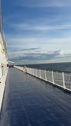 The video shows a deck of a ferry with a blue floor and a white railing. The sea and the sky are visible beyond the railing. The sun is shining brightly, reflecting on the water. There is a person standing near the railing looking out at the sea. The sky is blue with some clouds. The water is dark blue and appears calm with small waves.
