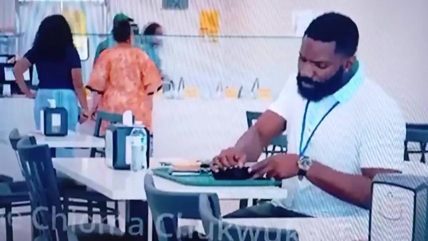 The video shows a man with a beard, wearing a light blue and white collared shirt and a lanyard, sitting at a table in a cafeteria. He is holding a black container and what appears to be a plastic fork or utensil. He is looking down at the food container and the utensil. In the background, there are other people standing in a line at a counter, and some are wearing masks. The cafeteria appears to have white tables and chairs, and a counter with food service equipment. The lighting is bright and appears to be from overhead fluorescent lights.