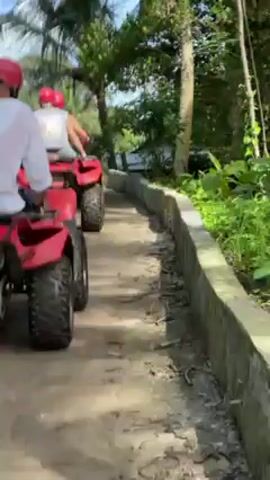 The video shows a group of people riding red ATVs on a dirt road. The riders are wearing red helmets and are traveling away from the camera. The road is lined with greenery and palm trees, suggesting a tropical environment. A building is visible in the background. The scene appears to be a guided ATV tour through a lush, possibly rural area.