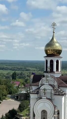 The video shows a close-up aerial view of a golden-domed Orthodox church. The dome is adorned with a cross and reflects the blue sky and white clouds. Below the dome, a belfry with arched openings is visible. The church itself is white with a brown roof. Scaffolding is present on the right side of the church, suggesting ongoing construction or renovation. In the background, a lush green forest stretches to the horizon under a partly cloudy sky. The scene is peaceful and serene, with no people or animals visible.