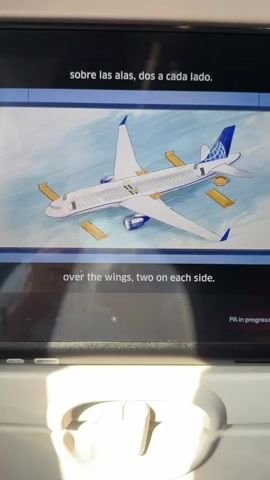The video displays a passenger's view of an in-flight entertainment screen showing an animated diagram of a United Airlines airplane. The screen displays safety information in both Spanish and English. The diagram illustrates the location of emergency exit ramp slides, with four slides shown inflated on the sides of the plane, two on each wing, and two at the rear exits. Text on the screen also indicates the presence of lifelines if needed. The camera captures the screen from a low angle, suggesting it's being viewed from a passenger seat. Part of the seat in front of the viewer is visible at the bottom of the frame, including a tray table latch. The ambient lighting suggests the cabin lights are on. A "PA in progress" indicator is visible on the screen.