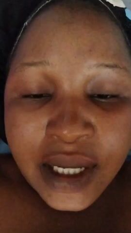 Here is a description of the video:
The video shows a person, likely a woman, close up from the chest up. She has dark skin, dark eyes, and dark lips. She is looking at the camera, blinking, and speaking. A black head covering with a white rim and bits of blue and white design is visible on her head. The background is blurred, with hints of blue fabric near her head.