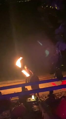 At night, a man is seen performing with two fire torches in an outdoor setting. He is wearing a dark shirt and dark pants. The torches are alight and emit significant light in the darkness. A low fence lined with lights separates the man from spectators sitting on beach chairs. One spectator is wearing a red shirt. The sand on the ground is visible around the seating. The lighting and torches create a festive atmosphere.