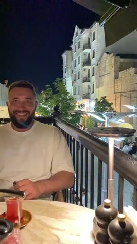 The video opens with a close-up of a man smiling and looking at the camera. He is wearing a light-colored t-shirt and sitting at a table. In the background, a balcony railing is visible, and beyond that, an outdoor seating area of a restaurant at night. People are seated at tables, and there is a hookah in the foreground. The camera then pans to the right, showing more of the outdoor restaurant area and the street beyond. Cars are parked along the street, and pedestrians are walking. Trees with green leaves are in the foreground, obscuring some of the view. The lighting is dim, with artificial lights illuminating the restaurant and street. The overall scene depicts a lively evening at an outdoor dining establishment.