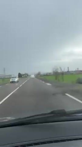 The video shows a car driving on a rural road on a cloudy day. The road is straight with a white line dividing the lanes. On either side of the road are fields, with some buildings and trees visible in the distance. The car's windshield wipers are in operation, suggesting rain or moisture on the windshield.