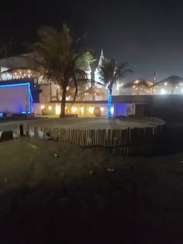 The video captures a nighttime view of an outdoor establishment, possibly a restaurant or resort, situated on a sandy area near a body of water. Several palm trees are visible, with their fronds illuminated by artificial lighting. The building features white walls and a thatched or similarly textured roof. Blue LED lights are strung along the exterior walls, adding a modern aesthetic. In the foreground, a low wooden fence encloses a portion of the area. The ground appears to be dark sand. The overall atmosphere is dimly lit, with the artificial lights creating pools of illumination against the dark night sky. There are no people visible in the video.