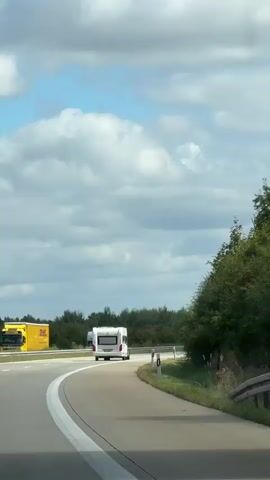 The video shows a view from inside a moving vehicle, looking forward and slightly to the right. The primary subject is a white travel trailer being towed on a highway. The trailer is in the right lane, moving away from the camera. In the background, to the left of the trailer, is a semi-truck with a gray trailer. Further in the distance, other vehicles and trees are visible. The road is paved with white lane markings. The environment appears to be rural or suburban, with trees lining the right side of the road and a cloudy sky above.