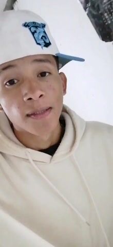 Here is a description of the video.

The video shows a young man in a beige hoodie and a white baseball cap with a blue brim. He is indoors, standing in front of a TV and some shelves. He is talking to the camera. The TV appears to be on. He is wearing a small earring in his left ear and a thin gold necklace.