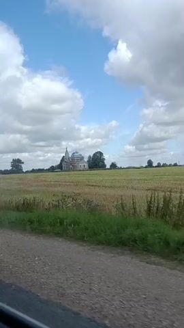 A church with blue domes is visible in the distance, situated in a field of dry grass under a partly cloudy sky. The foreground shows a dirt road and some green grass. The camera appears to be moving, suggesting it is being filmed from a vehicle.